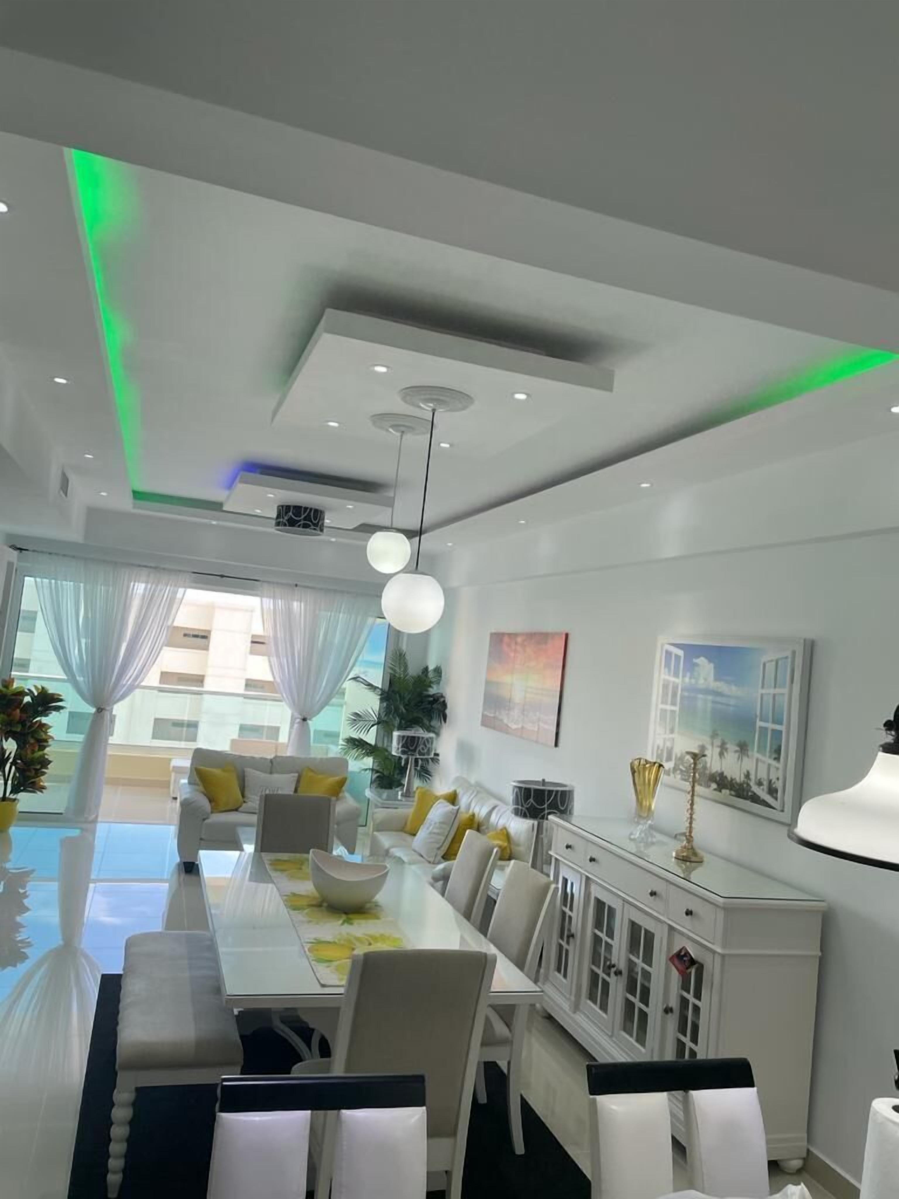 A modern dining area with a long table, white chairs, and a glass-top table. There are ceiling lights and a chandelier.