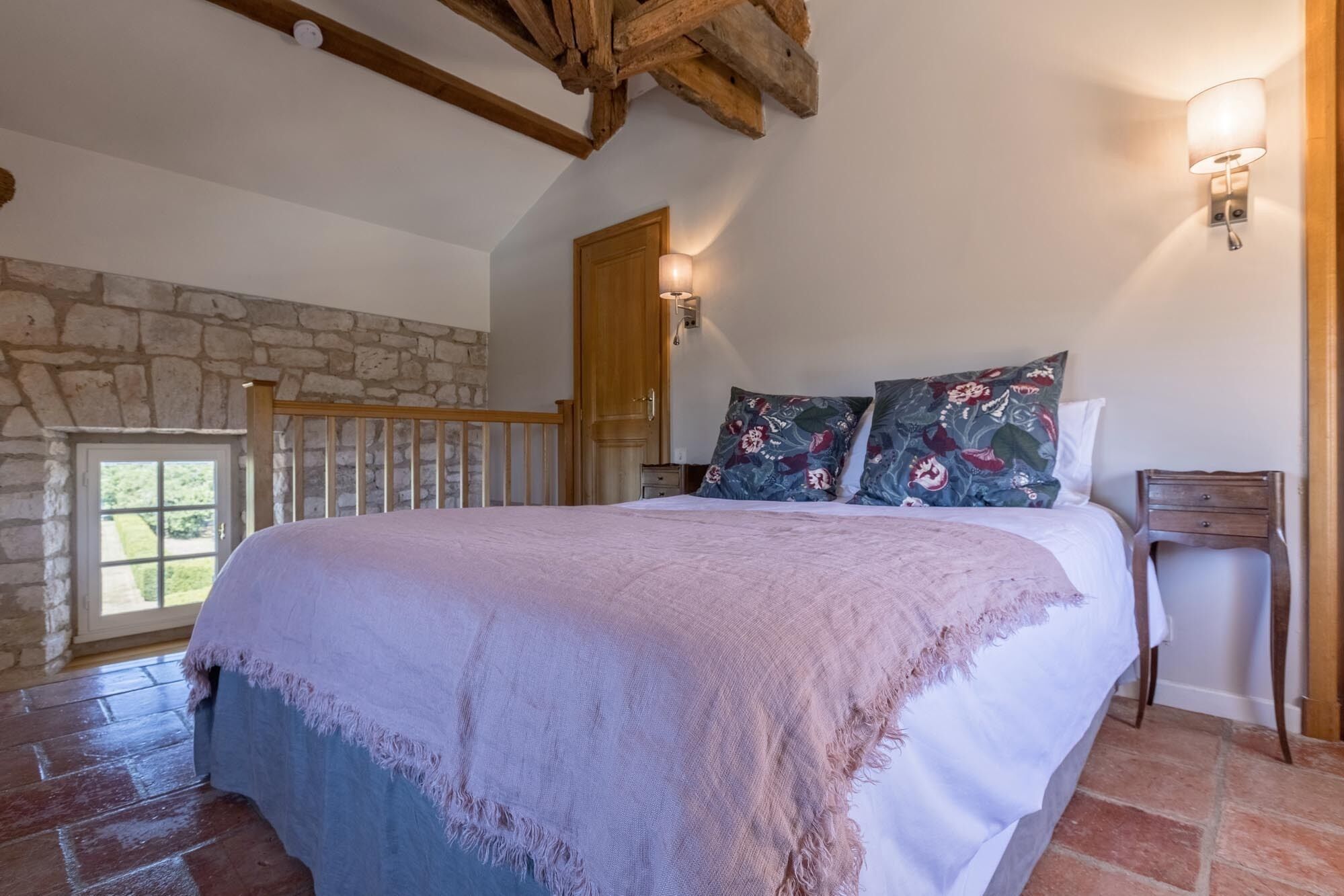 Une chambre avec un mur en pierre, une poutre en bois au plafond, un lit agrémenté de coussins à motifs floraux, une table de chevet et une fenêtre offrant une vue agréable.