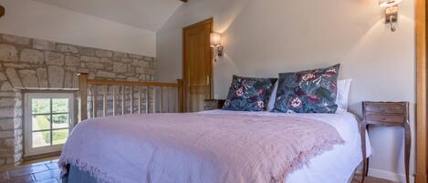 Une chambre avec un mur en pierre, une poutre en bois au plafond, un lit agrémenté de coussins à motifs floraux, une table de chevet et une fenêtre offrant une vue agréable.