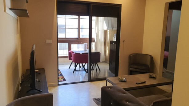 A living room with a sofa, a coffee table, and a TV. There is a sliding glass door leading to a balcony with a table and chairs. - Cosy studio. (Mansourieh)