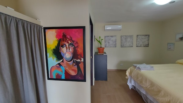 Une chambre d’hôtel avec un lit, un bureau agrémenté d’une plante en pot, un climatiseur et un tableau coloré accroché au mur.