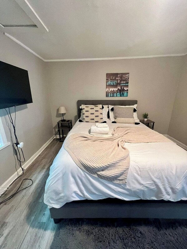 Un dormitorio ordenado con una cama grande, un televisor de pantalla plana en la pared, una mesita de noche pequeña con una lámpara, y una imagen enmarcada de un paisaje urbano sobre la cama.