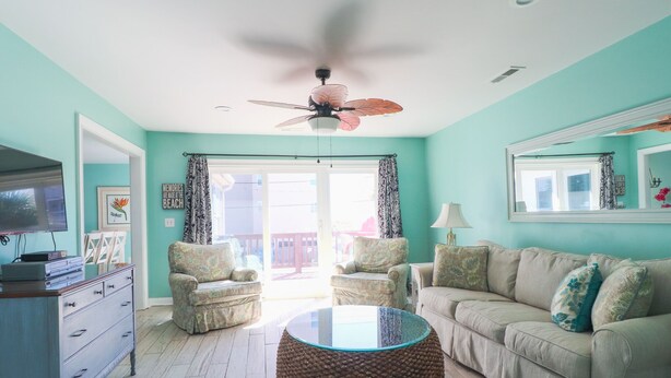 Un salon aux murs de couleur turquoise, doté d’un ventilateur de plafond, d’une table basse ronde en fibres tressées, d’un canapé beige et d’un téléviseur posé sur un meuble en bois.