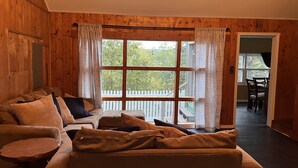 A cozy room with a wooden interior, a large window with curtains, a sofa, a small table, and a view of a white picket fence outside.