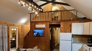 A loft-style room with a wooden ceiling, a television mounted on the wall, a kitchen area with white cabinets, and a dining area with a sofa and table.