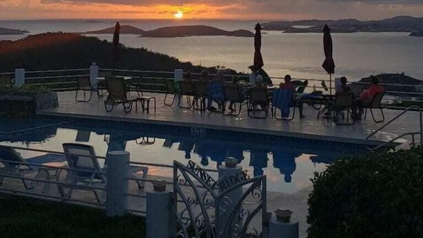 A pool area with lounge chairs and umbrellas, overlooking a body of water and distant hills during sunset.
