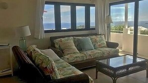 A living room with a sofa, a chair, and a glass coffee table. There are large windows with curtains, and a view of the outdoors.