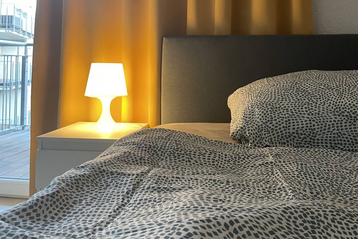 A hotel room with a bed, a nightstand with a lamp, and a view of the outside.