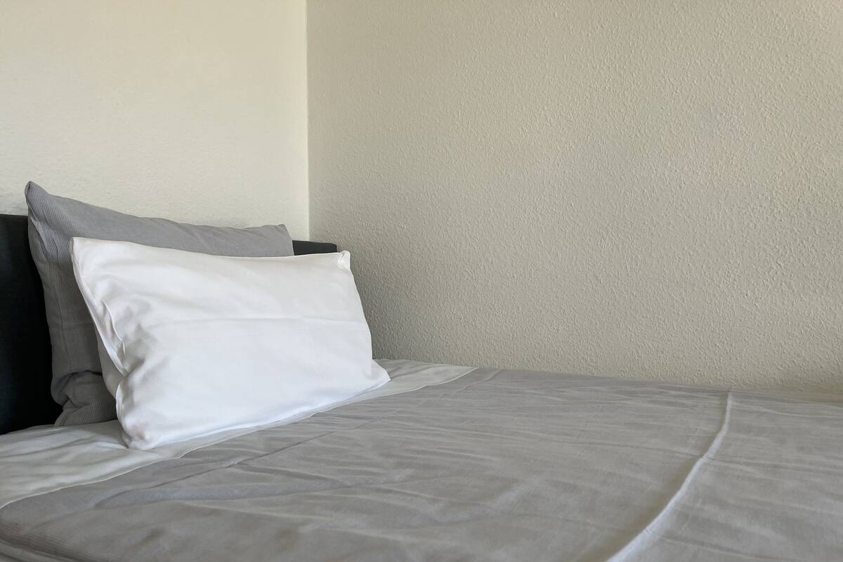 A neatly made bed with a grey bedspread and white pillows, a grey headboard, and a plain wall.