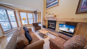 Ein gemütliches Wohnzimmer mit einer Couch, einem Sessel und einem Fernseher, der eine verschneite Berglandschaft zeigt.