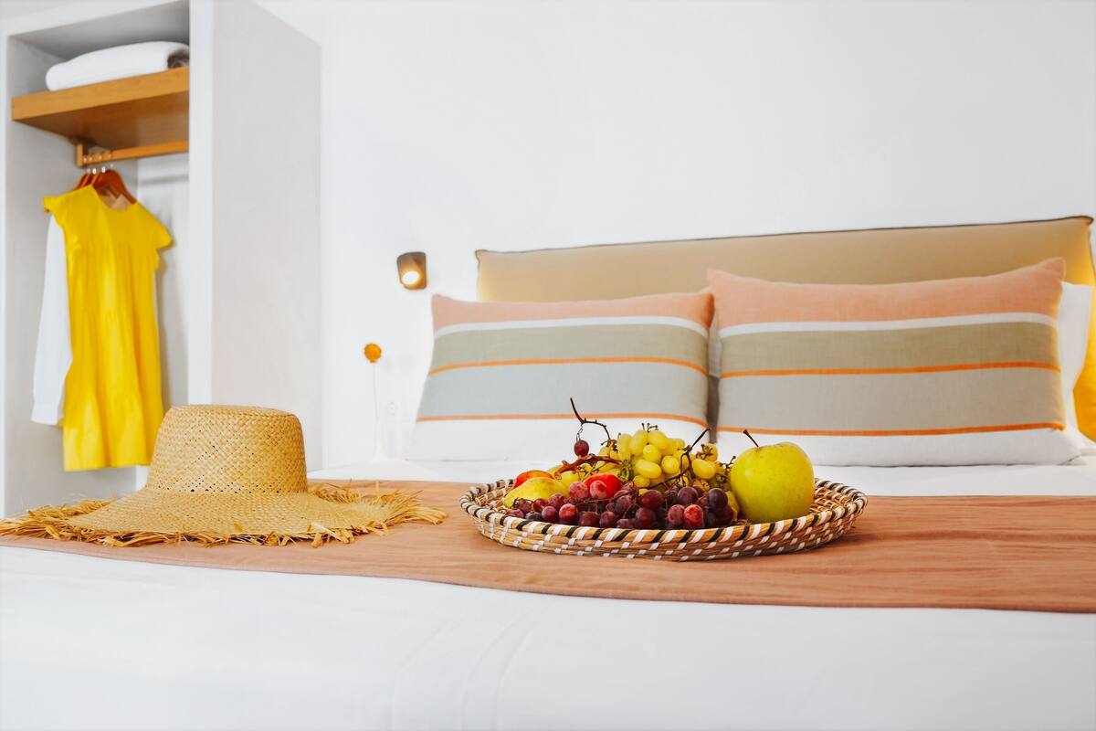 Una cesta de frutas ordenada sobre una cama, con un sombrero de paja a su lado, una prenda amarilla colgada en un perchero y una cama con almohadas a rayas.