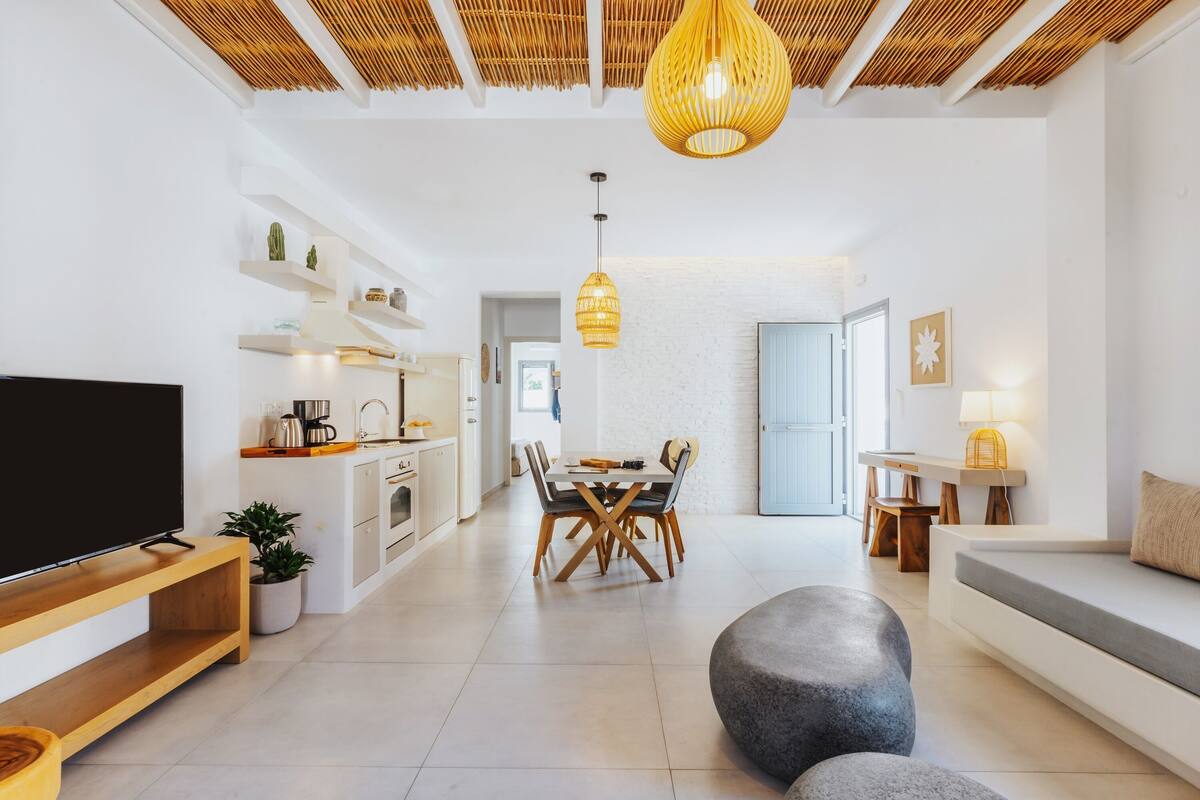 Una sala de estar moderna con televisor de pantalla plana, un comedor con mesa y sillas de madera, una cocina con armarios blancos y un techo con diseño tejido.