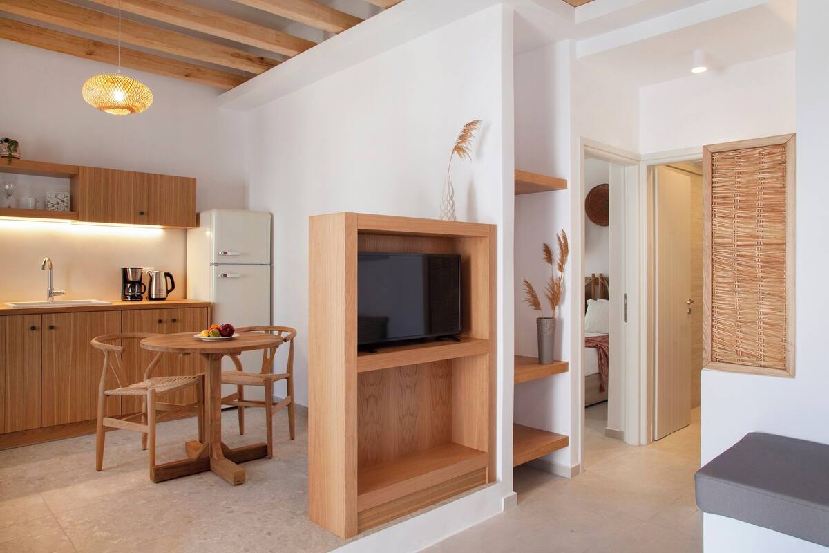 Una cocina moderna con armarios de madera, una mesa de comedor con sillas, televisión integrada y una pequeña zona de estar con un banco.
