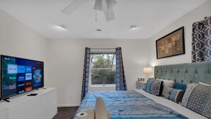 A bedroom with a bed, a TV on a stand, a window with curtains, and a framed picture on the wall.