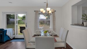 A dining area with a glass table, white chairs, a chandelier, and a view of the backyard through a sliding glass door.