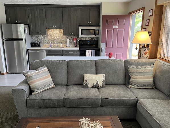 A living room with a grey sectional sofa, a wooden coffee table, and a kitchen area with dark cabinets, a stainless steel refrigerator, and a microwave.
