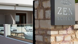 A stone wall with a sign for 'ZEN SUITES' and a glimpse of a modern building with a balcony and lounge chairs. - Zen play apartment,pool,Near beach & amenities (Nea Magnisia)