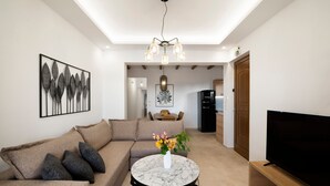 A modern living room with a sectional sofa, a round coffee table with a vase of flowers, a flat-screen TV, and a dining area in the background. - Zen play apartment,pool,Near beach & amenities (Nea Magnisia)