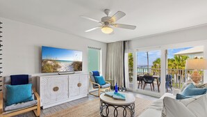 A modern living room with a ceiling fan, a large TV, a dining area with outdoor seating, and a view of the ocean. - MARAVILLA "LUCKY TOO" BEAUTIFUL PRIVATE BEACH VIEWS END UNIT "BEACH SETUP DAILY" (Miramar Beach)