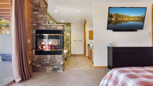 Une chambre avec une cheminée en pierre, un téléviseur fixé au mur et un lit recouvert d’une couverture à carreaux.