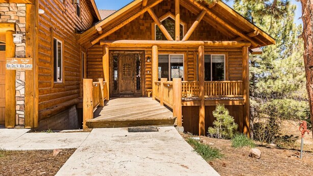 Une cabane en rondins dotée d’une terrasse en bois, de fondations en pierre et d’une enseigne indiquant qu’il s’agit de la « Sky High Cabin », pouvant accueillir un maximum de 14 personnes et où l’on peut garer un maximum de 6 voitures.