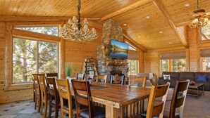 Un intérieur spacieux, avec des éléments en bois, comprenant une grande table à manger entourée de chaises, une cheminée en pierre et un téléviseur fixé au mur.