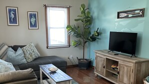 A living room with a sofa, a coffee table, a TV on a wooden cabinet, and a window with blinds. - On a Whim, a family retreat! (Chincoteague)