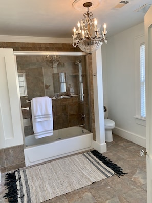 A modern bathroom with a glass-enclosed shower, a white bathtub, a chandelier, and a white toilet.