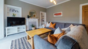 A living room with a grey sofa, a wooden coffee table, a flat-screen TV on a white stand, a fireplace, and a bookshelf. - The Riverside Apartment (free parking) (Stirling)