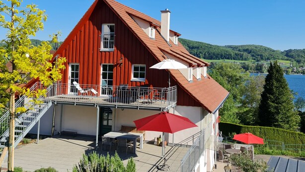 A red house with a balcony, a pool, and a view of a lake and hills.