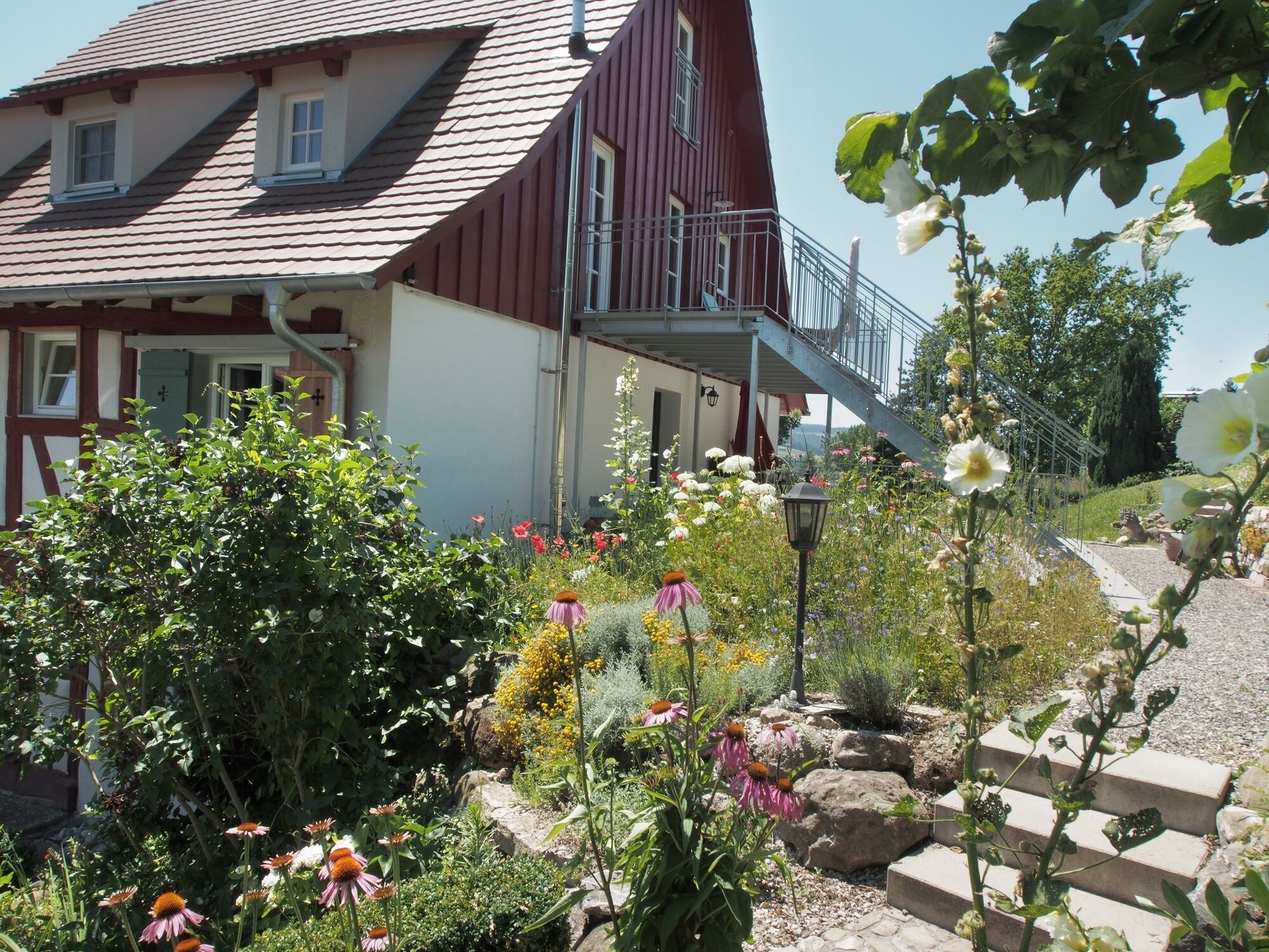 Une maison traditionnelle avec un toit rouge et des murs blancs, entourée d’un jardin bien entretenu où poussent différentes sortes de fleurs et de plantes.