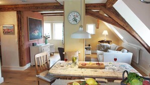A cozy loft apartment with exposed wooden beams, a dining area with a wooden table set for a meal, and a living area with a sofa and a lamp.