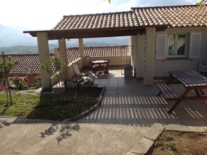 A covered patio with a table and chairs, a garden area with plants, and a view of the mountains in the background. - House (Poggio-d'Oletta)