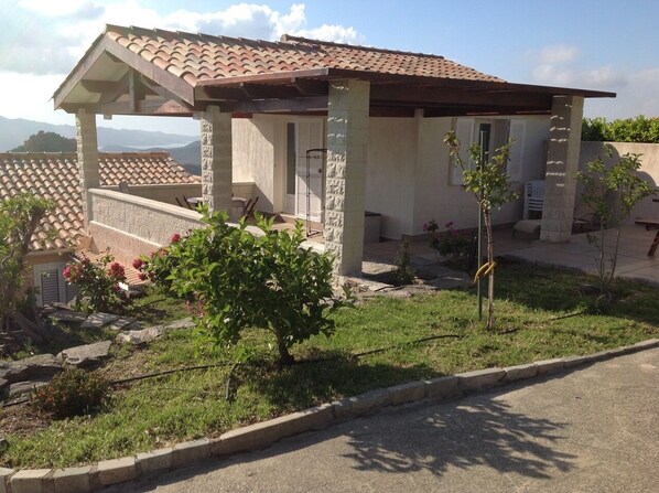 A house with a tiled roof, a covered patio, and a small garden with a tree and flowers. - House (Poggio-d'Oletta)
