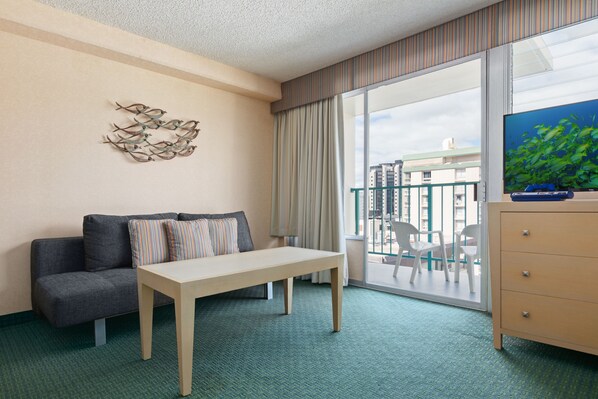 Ein Hotelzimmer mit einer Couch, einem Tisch, einem Fernseher, einem Schminktisch und einem Balkon mit Blick auf Gebäude.