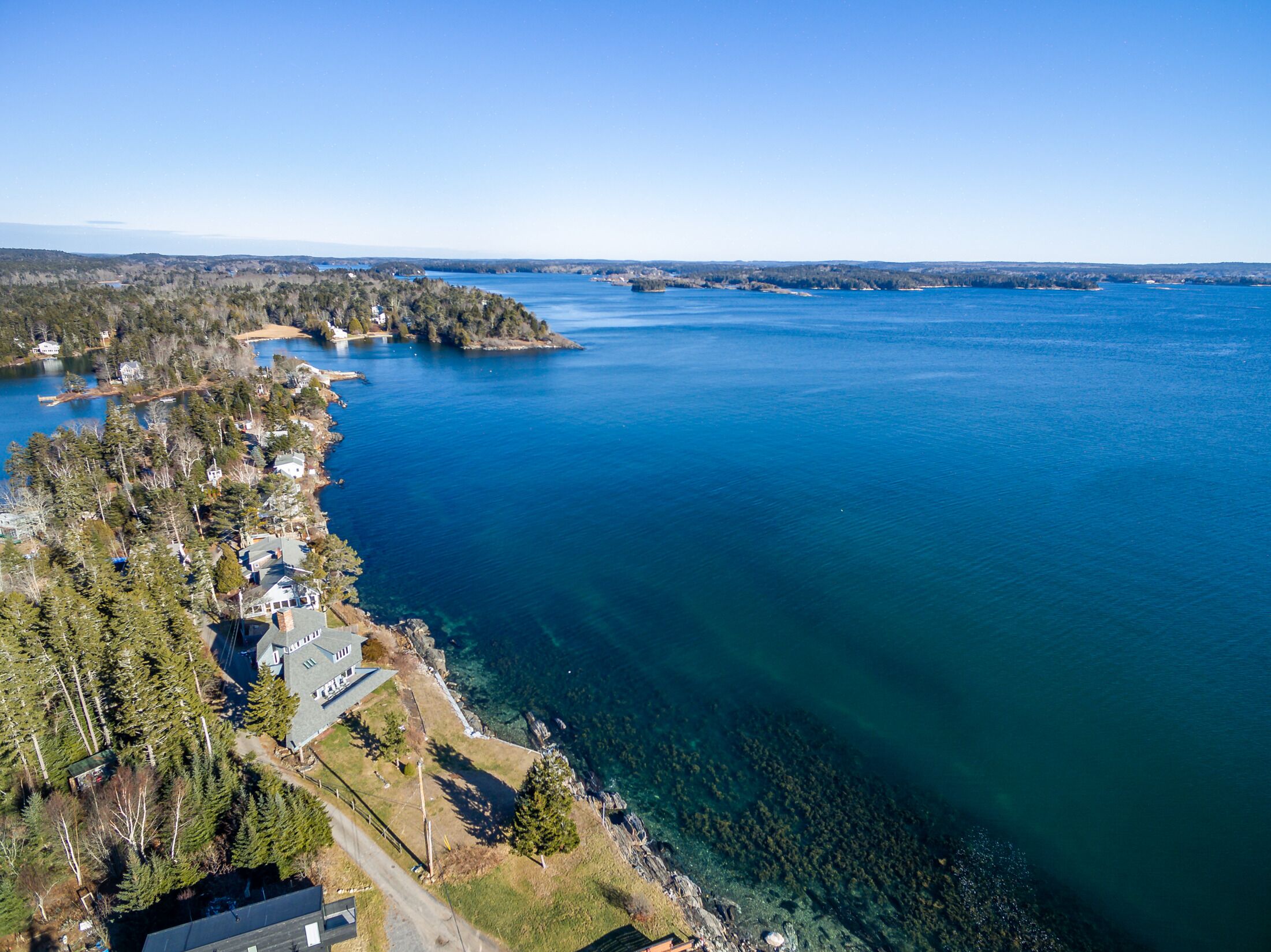 An aerial view of a coastal property with houses along the shoreline, clear blue water, and a forested area.