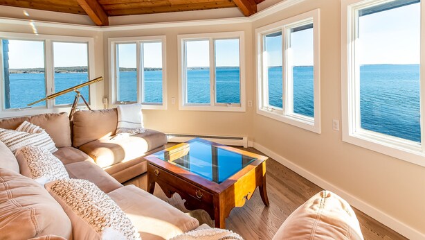 A room with a large window overlooking the sea, a wooden ceiling, and a glass-top coffee table. - Sunrise Catcher with a Panoramic View!  (Harpswell)