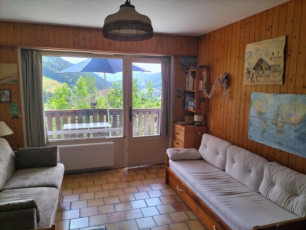 A cozy room with a wooden interior, a sofa, a chair, a large window with a view of mountains, and a globe on the wall. - Beautiful mountain views from the studio. Gondola lift at 500 m.  Free shuttle bus  (Le Grand-Bornand)