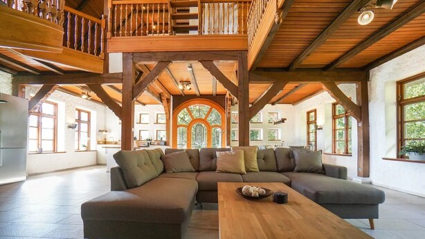 Een ruime woonkamer met een grote modulaire bank, een houten salontafel en een hoog houten plafond met zichtbare balken.