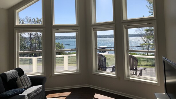 A room with large windows offering a view of a lake and dock. There is a ceiling fan, a couch, and a flat-screen TV. - Waterfront Lake House - Portage Lake - 4 Beds, 3 Baths, Private Dock, Kayaks (Onekama)