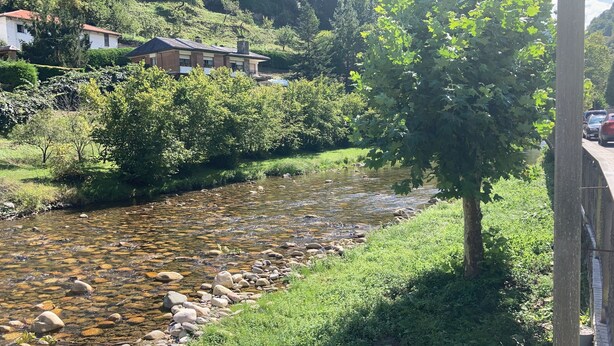 A river with clear water and rocks, surrounded by greenery and a building with a red roof.
