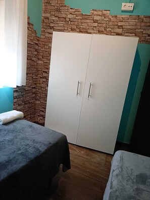 A room with a wardrobe, two beds, a window with curtains, and a wall with a stone-like texture.