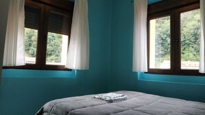 A room with teal walls, two windows with white curtains, and a bed with a grey quilt.