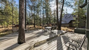 Une terrasse en bois, agréée de mobilier d’extérieur, entourée d’arbres et bordée d’un bâtiment doté d’un toit en pente.