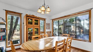 Un espace repas comprenant une table et des chaises en bois, un buffet en bois doté de portes en verre, et de grandes fenêtres offrant une vue sur l’extérieur.