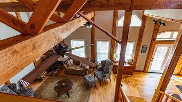 A spacious loft-style room with a high ceiling, exposed wooden beams, a ceiling fan, a sofa, a chair, and a small table.