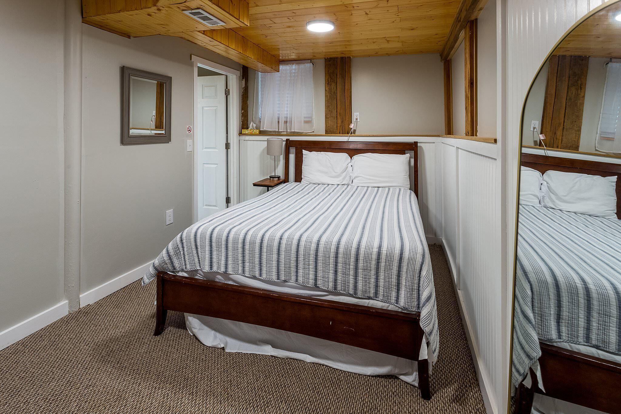 A bedroom with a wooden bed frame, a white door, a small bedside table with a lamp, and a window with a curtain.