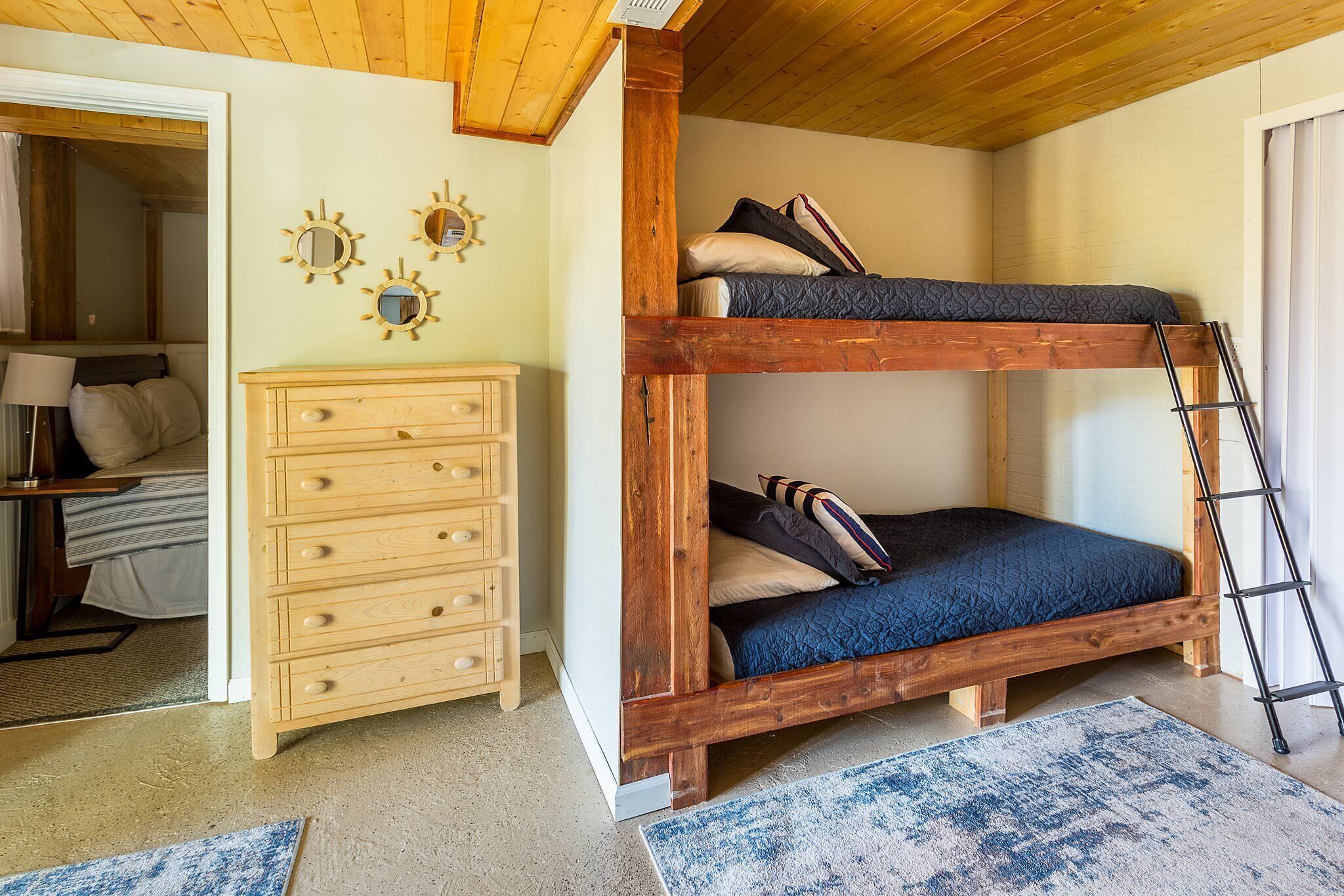 A bunk bed with a ladder, a wooden dresser, and a small rug in a room with wooden ceiling beams.