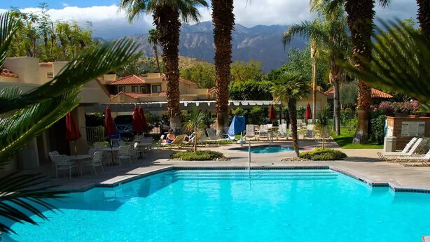 Une piscine entourée de chaises longues et de parasols, bordée de palmiers et avec une chaîne de montagnes en toile de fond.
