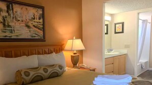 Une chambre d’hôtel avec un lit, un tableau, une lampe, une salle de bain avec un lavabo et des serviettes sur le sol.